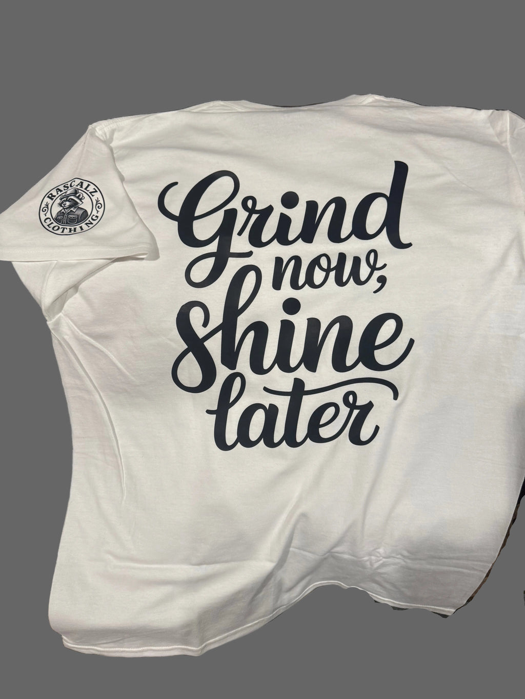 White tank top with 'Grind now, shine later' text on a wooden surface.