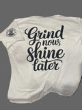 White tank top with 'Grind now, shine later' text on a wooden surface.